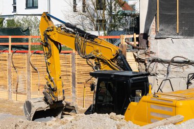 Ağır makineler aktif olarak kalabalık bir kentsel alandaki bir inşaat alanında kazı yapıyor. Kazıcı temiz mavi gökyüzü altında toprak ve materyalleri taşırken, işçiler projeyi yönetiyor.