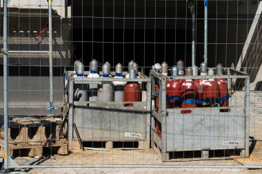 Çeşitli gaz tüplerinden oluşan bir koleksiyon, bir inşaat alanındaki çitli bir alanda metal kafeslerde güvenli bir şekilde depolanır. Titiz düzenleme, sanayi ortamlarındaki güvenlik tedbirlerini vurguluyor.