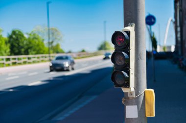 Kırmızı ışıklı bir trafik lambası bir arabanın geçtiği yolun yanında duruyor. Hava güneşli ve açık, bölgeyi aydınlatıyor ve caddeyi çevreleyen yeşil ağaçlar....
