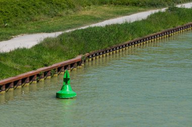 Sakin sularda yüzen yeşil bir şamandıra manzaralı bir su yolundaki navigasyon yollarını gösteriyor. Bereketli yeşillik ve bakımlı bir kıyı şeridi bu güneşli günde huzurlu atmosferi güçlendirir..