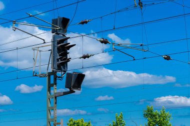 Bir demiryolu sinyali kabarık bulutlar ve açık mavi bir gökyüzü karşısında dimdik ayakta duruyor. Elektrik hatları gerilirken sinyal renklerini gösterir, bu da iyi korunmuş bir demiryolu ağını gösterir.