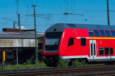 Modern bir kırmızı tren, yeşil çimenler ve tren raylarıyla çevrili, açık mavi bir gökyüzü altında istasyona yaklaşıyor. Elektrik hatları ve kulübe olay yerinin endüstriyel zeminini oluşturuyor..