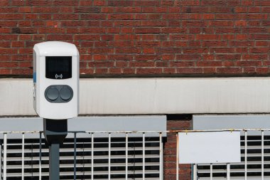 Modern bir parkmetre kırmızı tuğla bir binanın önündeki direğe monte edilir. Belgede, kentsel park çözümleri için ileri teknolojiyi gösteren dijital bir ekran ve ödeme düğmeleri yer alıyor..