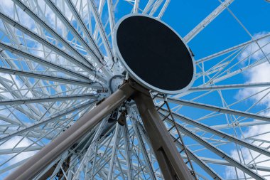 Görüntü, telleri ve destek kirişlerini gösteren bir dönme dolabın karmaşık çerçevesini yakalıyor. Güneş ışığı bulutların arasından süzülür, gündüz vakti parlak ve davetkar bir atmosfer yaratır..