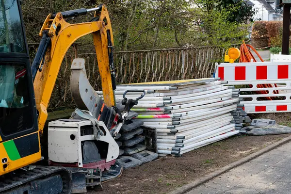 Bir inşaat sahasında, bariyerler ve destek de dahil olmak üzere düzgünce istiflenmiş inşaat malzemelerinin yanında bir kazıcı sergileniyor. Atmosfer sakin, tipik bir çalışma gününü yansıtıyor..