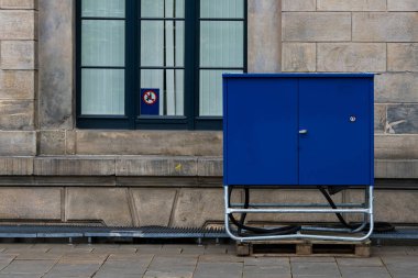 Yakınlarda büyük bir penceresi olan eski bir taş duvarın yanında parlak mavi bir servis kutusu duruyor. Sahne, şehir ortamında modern altyapı ve tarihi mimarinin bir karışımını taşıyor..