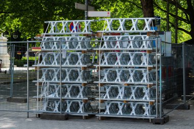 Metal bariyerler, halka açık bir parktaki çitlerle çevrili bir alanda düzgünce istiflenmiş. Parlak güneş ışığı ağaçların arasından süzülerek çevredeki sakin atmosferi arttırır..