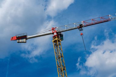 Uzun bir vinç, tüylü beyaz bulutlarla dolu parlak mavi bir gökyüzüne karşı belirgin bir şekilde duruyor. Canlı kırmızı ve sarı renkleri, şehir gelişimini sembolize ediyor..