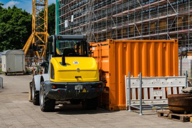 Sarı ve gri bir forklift, bir inşaat alanında turuncu bir nakliye konteynırının yanına yerleştiriliyor. Bina iskelesi ve yapı malzemeleri arka planda berrak mavi bir gökyüzünün altında görünür..