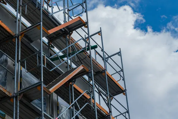 İskeleler, çeşitli seviyeler ve platformlar sergilenen bir inşaat alanında dimdik ayakta duruyor. Parlak mavi gökyüzü ve kabarık bulutlar endüstriyel yapıya çarpıcı bir kontrast sağlar..
