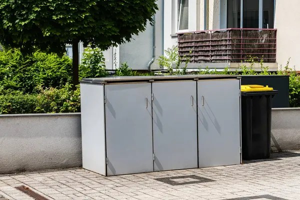 Üç panelli depolama ünitesi kentsel alanda bir geri dönüşüm kutusunun yanında duruyor. Yeşillik ve apartman arkaplanı oluşturur. Gündüz gözüyle temiz bir ortam sunar..