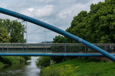 Çağdaş bir köprü, yemyeşil ve ağaçlarla çevrili sakin bir nehir boyunca uzanır. Bulutlu gökyüzü, suyun kenarındaki huzurlu şehir manzarası için sakin bir zemin oluşturur..