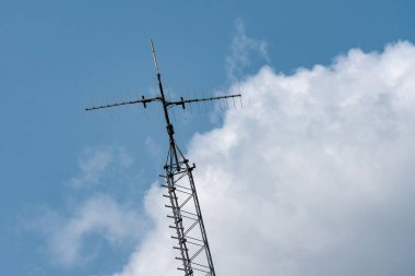 Uzun bir iletişim kulesi, tüylü beyaz bulutlarla benekli, parlak mavi bir gökyüzüne doğru yükseliyor. Yapı sinyal iletimi için antenlerle donatılmıştır.