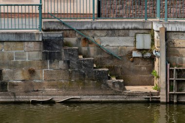 Metal bir korkulukla çevrili yıpranmış taş bir merdiven durgun suya iner. Aşırı büyümüş bitkiler, kenar mahallelere tutunarak, deniz kenarlarının huzurunu yansıtıyorlar..
