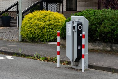 Otoyoldaki elektrikli araç şarj istasyonu, güvenlik için kırmızı ve beyaz direklerle işaretlenmiş. Yakınlarda, dingin yeşil bir çalı sakin bir günde kentsel alana biraz doğa katar..