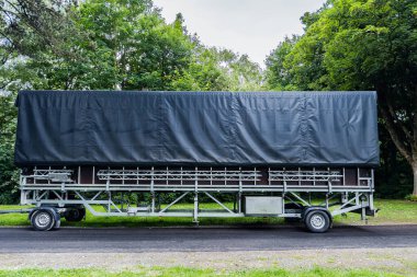 Üzerinde siyah muşamba kapağı olan büyük ve ağır yük taşıyan bir karavan bir ağaç hattının yanında park halinde. Çevrede ağaçların gölgesi ve yeşilliği var. Bu da açık havada sakin bir ortam olduğunu gösteriyor..
