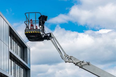 Mavi gökyüzü ve açık bir günde dağınık bulutlarla çevrili modern bir binanın yanında çelik bir kabin bulunan katlanmış hidrolik vinç. Güvenlik konileri asansör platformunda görünür..