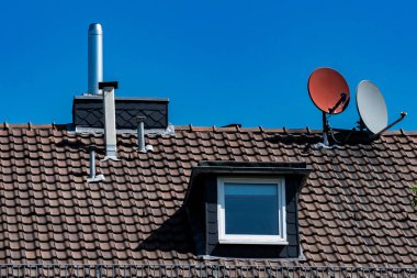 Parlak güneş ışığı, uydu antenleri ve bacası olan bir çatıya vurgu yapıyor. Açık mavi gökyüzü, çatının eşsiz özelliklerini ve dokularını vurgulayan bir zemin görevi görür..