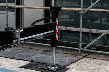 İnşaat ekipmanları bir şehir caddesinde, iskele ile çevrili. Kurulumda güvenlik işaretleri olan metal bir direk ve sağlam bir taban yer alıyor. Bu da yenileme çalışmalarının devam ettiğini gösteriyor..