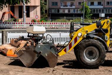 Şehir ortamında bir inşaat alanında çalışan sarı bir kazıcı görülüyor. Makine zemini kazarken diğer ekipmanlar arka planda berrak mavi gökyüzünün altında görünüyor..