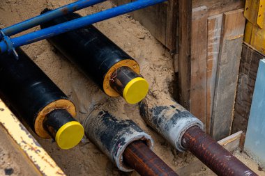 Construction workers are installing utility pipes in a deep trench filled with sand. The pipes are secured with wooden supports, indicating a significant underground infrastructure upgrade.