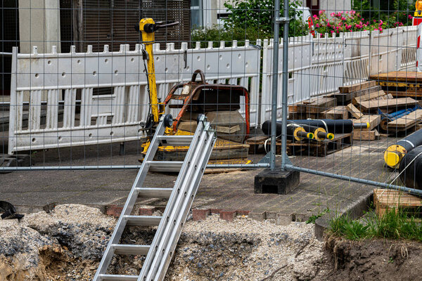 A construction site features a ladder leaning against a fenced area. Nearby, various equipment and materials are organized, indicating ongoing work in the urban location during daylight hours.