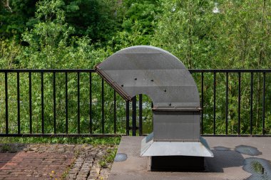 Bereketli yeşillik tarafından çerçevelenmiş sakin bir su kütlesinin yanında eşsiz bir metal havalandırma yapısı bulunur. Güneş ışığı manzarayı aydınlatır, açık hava ortamının sakin atmosferini arttırır..