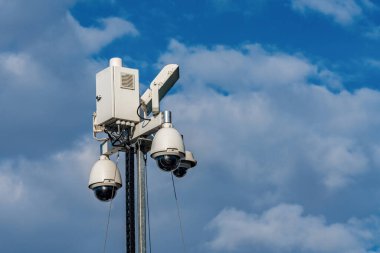 Uzun bir direğe yerleştirilmiş bir güvenlik kamerası sistemi çevreyi gözetliyor, birden fazla kamera birimiyle donatılmış. Parlak bulutlar mavi gökyüzüne karşı arkaplan oluşturur, bu da gündüz vakti.