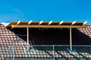 Bir yerleşim yerindeki eğimli bir çatıda ahşap bir iskelet inşa ediliyor. Mavi gökyüzü, iskele ve inşaat malzemeleri yakınlarda görünürken bir zemin görevi görüyor..