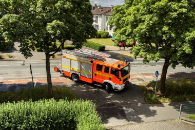 Parlak turuncu bir itfaiye aracı yemyeşil ağaçlarla çevrili bir şehir caddesinde park halinde. Kamyon harekete hazır, yakınlardaki binalar ise gün ışığında kentsel bir zemin sağlıyor..