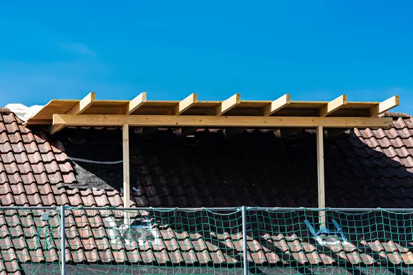 Bir yerleşim yerindeki eğimli bir çatıda ahşap bir iskelet inşa ediliyor. Mavi gökyüzü, iskele ve inşaat malzemeleri yakınlarda görünürken bir zemin görevi görüyor..