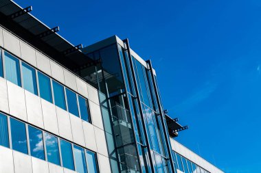 Çağdaş bir ofis yapısında güneş ışığını yansıtan parlak cam bir cephe bulunur. Tasarım temiz çizgileri vurguluyor ve açık mavi gökyüzüne karşı canlı bir zemin sunuyor.