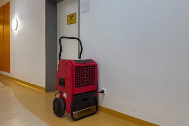 Koridordaki bir duvarda kırmızı bir nemlendirici var. Yakınlarda bir tuvalet olduğunu gösteren bir tabela var. Alan düz zemin ve temiz, basit çizgiler içeriyor, fonksiyonel bir atmosfer yaratıyor..