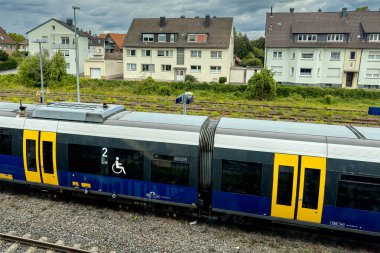A modern train waits at a quiet suburban station surrounded by residential buildings. The atmosphere feels calm as clouds hover overhead during afternoon hours.