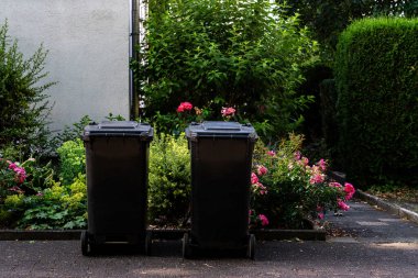 İki siyah çöp bidonu canlı çiçekler ve yeşilliklerle çevrili bir yolda duruyor. Sahne huzurlu bir yerleşim bölgesinde geçiyor, doğa ve günlük yaşamın bir karışımını sergiliyor..