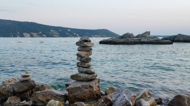 Karadağ 'ın Herceg Novi kentindeki Kotor Körfezi' nin kayalık kıyısında dikkatlice dengelenmiş taş yığınlarının (cairns) yer aldığı sakin bir manzara. Koyun sakin ve berrak suları uzak dağlara doğru uzanıyor ve huzurlu ve meditasyon yapan bir atmosfer yaratıyor.