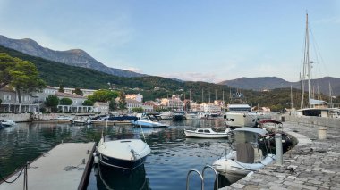 Karadağ 'ın Herceg Novi kentinde sakin ve berrak sularda sallanan çeşitli yat ve teknelerle dolu bir marina manzarası. Sahne, parlak bir gökyüzünün altında yuvarlanan yeşil dağların zeminine kurulur..