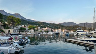 Karadağ 'ın Herceg Novi kentinde sakin ve berrak sularda sallanan çeşitli yat ve teknelerle dolu bir marina manzarası. Sahne, parlak bir gökyüzünün altında yuvarlanan yeşil dağların zeminine kurulur..
