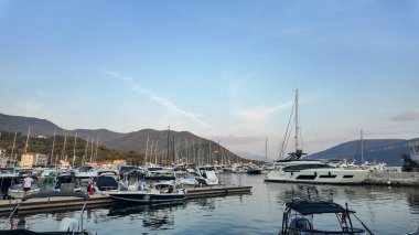 Karadağ 'ın Herceg Novi kentinde sakin ve berrak sularda sallanan çeşitli yat ve teknelerle dolu bir marina manzarası. Sahne, parlak bir gökyüzünün altında yuvarlanan yeşil dağların zeminine kurulur..