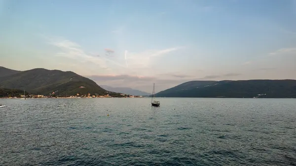 Karadağ 'ın Herceg Novi kentindeki Kotor Körfezi' nin sakin sularında süzülen yalnız bir yelkenlinin huzurlu bir görüntüsü. Göz kamaştırıcı arka plan, suyun kenarından yükselen görkemli dağları resmedilmeye değer ve sakin bir manzara yaratıyor.. 