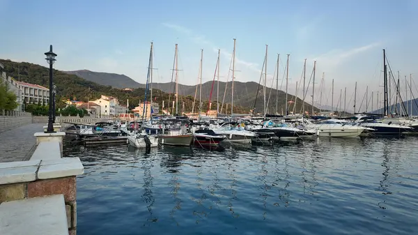 Karadağ 'ın Herceg Novi kentinde sakin ve berrak sularda sallanan çeşitli yat ve teknelerle dolu bir marina manzarası. Sahne, parlak bir gökyüzünün altında yuvarlanan yeşil dağların zeminine kurulur..