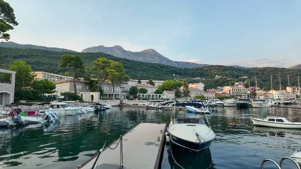 Karadağ 'ın Herceg Novi kentinde sakin ve berrak sularda sallanan çeşitli yat ve teknelerle dolu bir marina manzarası. Sahne, parlak bir gökyüzünün altında yuvarlanan yeşil dağların zeminine kurulur..