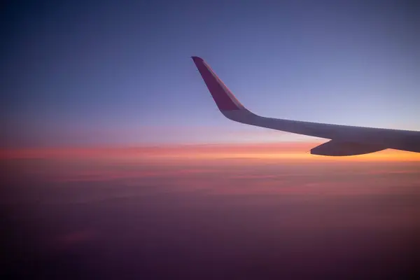 Bir uçak kanadı çarpıcı bir alacakaranlık gökyüzüne karşı yakalanır. Mavi mor turuncu ve pembe bir gradyan, hava yolculuğunu ve bulutların üzerinden gün batımının dingin güzelliğini simgeler..