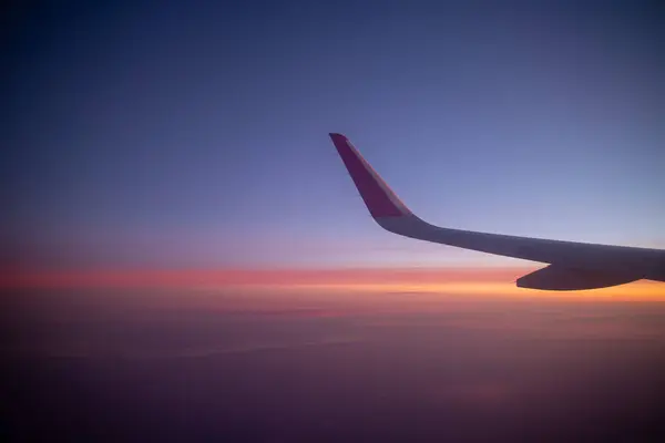 Bir uçak kanadı çarpıcı bir alacakaranlık gökyüzüne karşı yakalanır. Mavi mor turuncu ve pembe bir gradyan, hava yolculuğunu ve bulutların üzerinden gün batımının dingin güzelliğini simgeler..