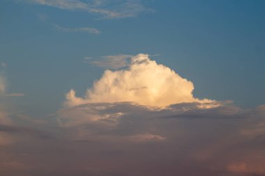 Çarpıcı bir kümülonimbus bulutu gökyüzünde belirgin bir şekilde duruyor. Güneş batarken maviden yumuşak pembe renklere geçiş yapıyor. Canlı ve dramatik bir atmosfer manzarası çiziyor..