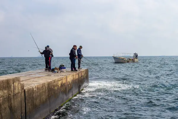 Bir grup insan, Odesa Ukrayna 'da sağlam bir beton iskelede balık tutarken görülüyor. Yapıya çarpan dalgalar ve Karadeniz' de uzak bir balıkçı teknesi, tipik bir kıyı sahnesi ve popüler eğlenceyi resmediyor..