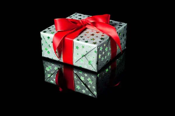 Single green gift box with red ribbon bow on black reflective surface. Gift box wrapped in green shine paper with red ribbon