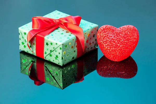 Single green gift box with red heart on blue reflective surface. Gift box wrapped in green shine paper with red ribbon