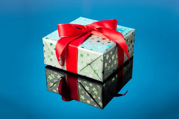 Single green gift box with red ribbon bow on blue reflective surface. Gift box wrapped in green shine paper with red ribbon close up