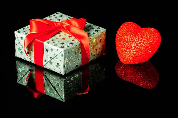 Single green gift box with red heart on black reflective surface. Gift box wrapped in green shine paper with red ribbon